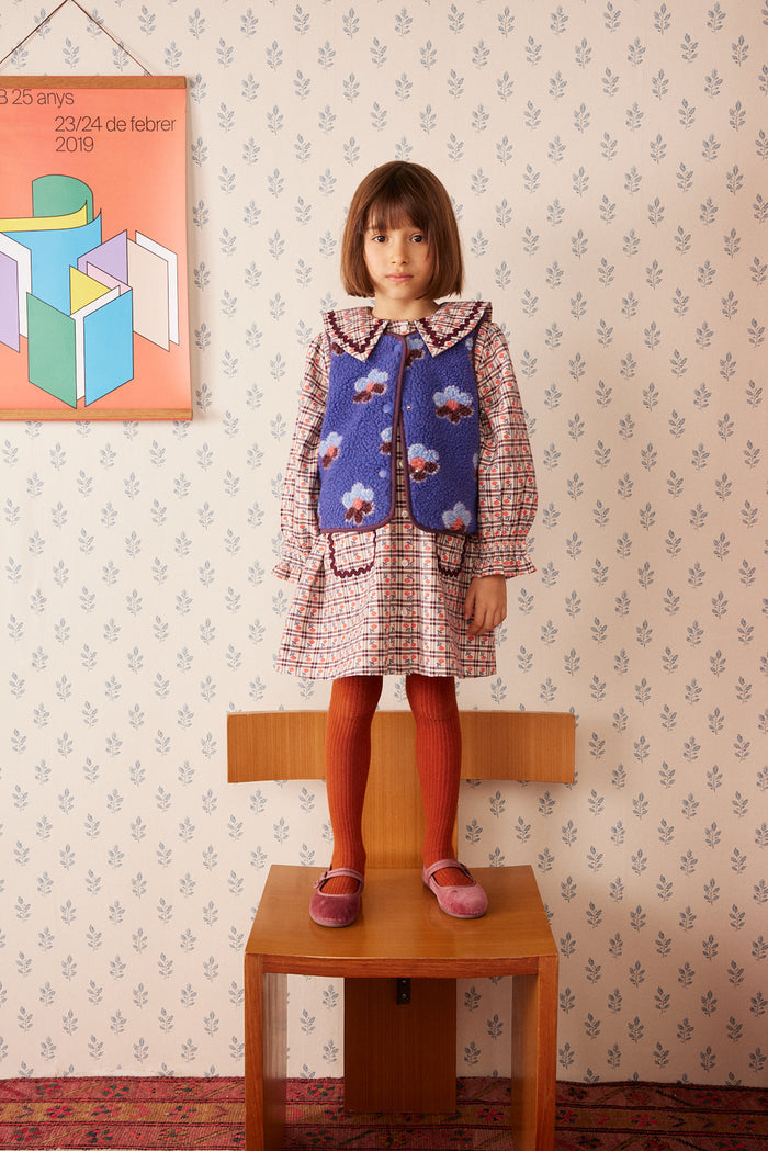 A young girl with brown hair is wearing a vintage-style plaid dress with a blue floral fleece vest, and red tights.