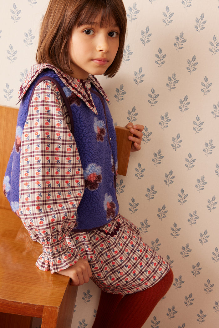 A young girl with brown hair is wearing a vintage-style plaid dress with a blue floral fleece vest, red tights, and a matching plaid bow headband. She is leaning against a wooden chair in front of a beige wallpapered wall with a blue leaf pattern.