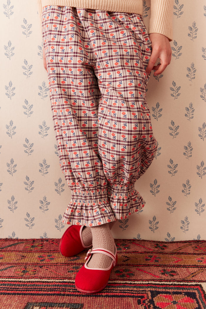 A person wearing patterned pleated trousers with a red and beige floral plaid design, paired with red Mary Jane shoes and socks, standing on a patterned rug against a floral wallpaper background.