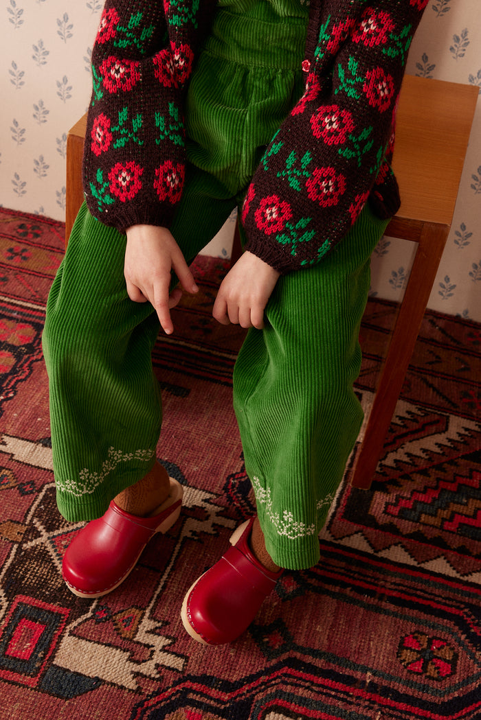 A child seated on a wooden chair wearing bright green corduroy pants, a dark knit sweater with red and pink floral patterns, and red clogs on a patterned rug.