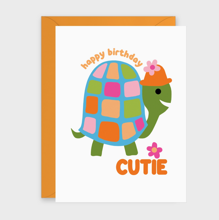 A birthday card with an orange envelope behind it, featuring a cartoon turtle with a multicolored patchwork shell, green head and legs, an orange hat with a pink flower, and the words 'happy birthday' and 'CUTIE'.