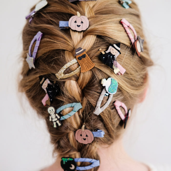A close-up of a person's styled blonde braided hair adorned with multiple Halloween-themed hair clips, including pumpkins, a witch, a skeleton, and a ghost.