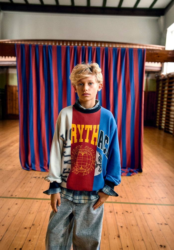 Person wearing a colorful sweater with text and graphics in an indoor setting with red and blue curtains.