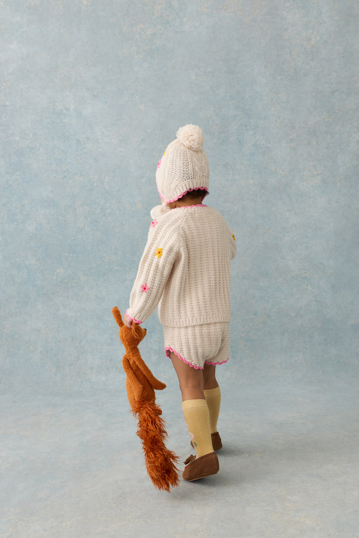 Child wearing a white knit outfit with a fluffy hat, holding a toy, against a light blue background