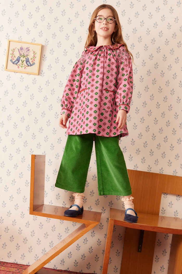 A young girl with glasses stands on wooden steps wearing a pink patterned long-sleeve blouse with a ruffled collar and green wide-leg pants, set against a vintage floral wallpaper background.