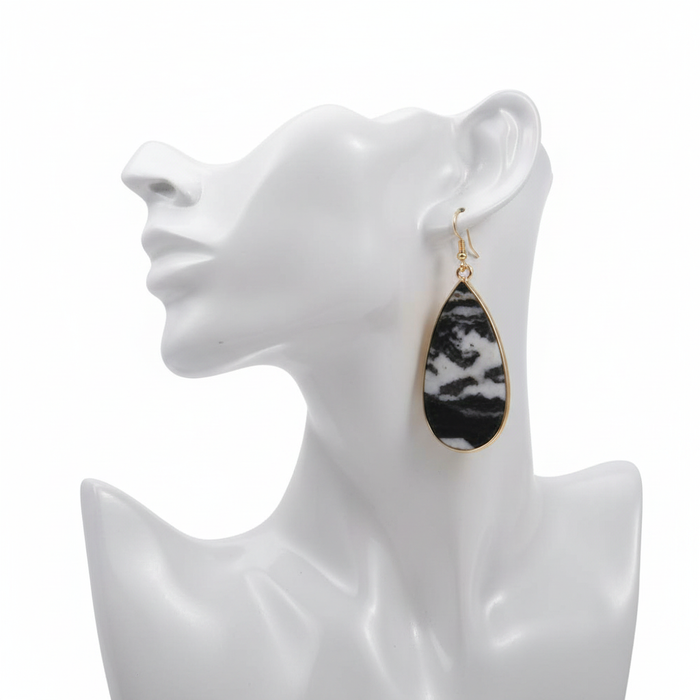 A single statement dangle earring featuring a large teardrop-shaped zebra jasper stone with natural black and white marbled patterns. The stone is encased in a thin gold-toned metal frame and hangs from a simple gold French hook. It is displayed on a white mannequin head against a clean white background.