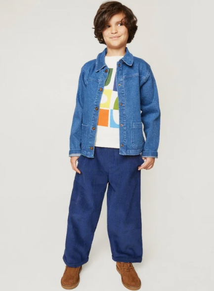 A child model wearing a blue denim jacket, blue corduroy pants, a white graphic t-shirt, and brown suede boots, standing against a light gray studio background.