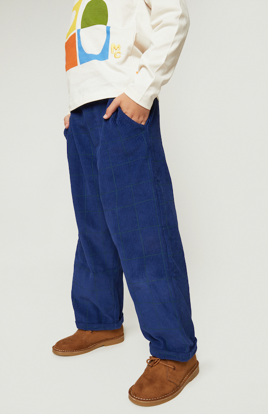 A person wearing loose blue corduroy trousers with a subtle windowpane check, styled with a cream sweatshirt and brown suede shoes against a light gray studio background.