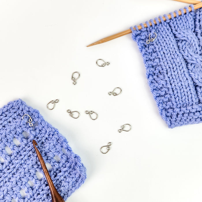 Cast melody clip stitch markers scattered on white background between a blue crochet swatch with hook and a blue knit swatch on bamboo needle, showing how the clips attach to both crafts