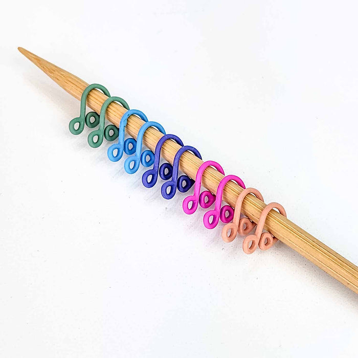 Row of pastel rainbow Cast Melody Clip locking stitch markers in pink, purple, blue, green, and yellow lined up on a bamboo knitting needle