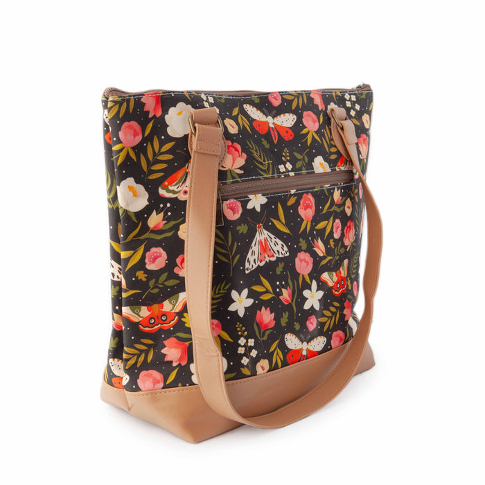 Night Forest Mega Trinity Bag three-quarter view showing the side gusset, zippered exterior pocket, and tan vegan leather shoulder strap with the dark floral and moth print
