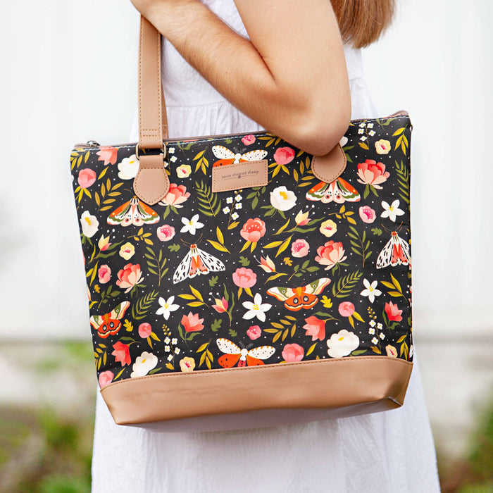 Person carrying the Night Forest Mega Trinity Bag on one shoulder showing the dark floral and moth print canvas with tan vegan leather bottom and strap against a white dress