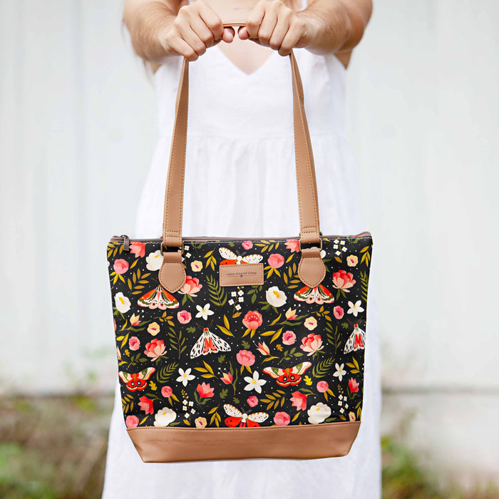 Person holding the Night Forest Mega Trinity Bag by both handles showing the full front with the dark floral and moth print canvas, Twice Sheared Sheep label, and tan vegan leather bottom
