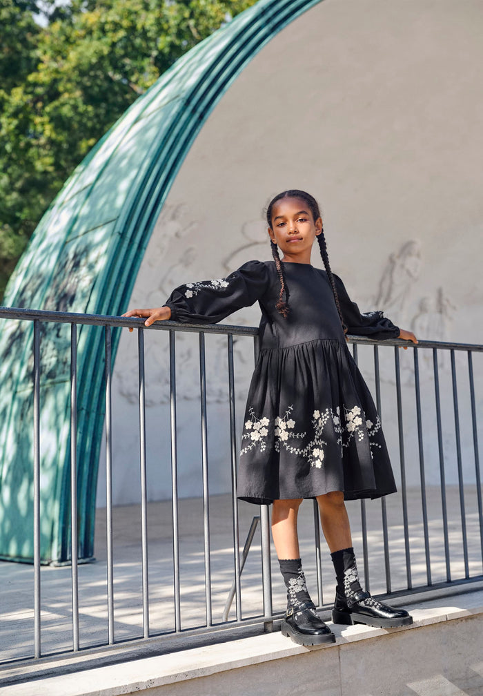 Young girl in a black dress with floral patterns standing on a balcony.