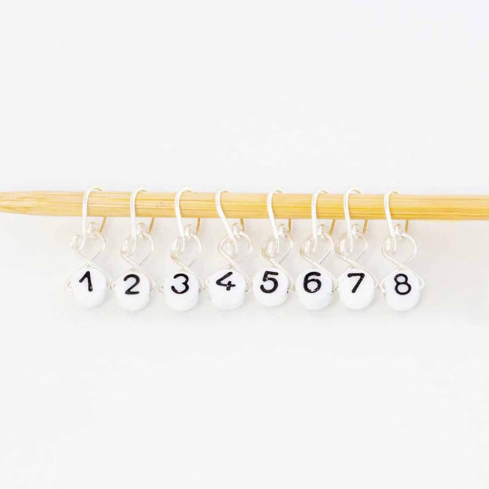 Silver melody clip style numbered stitch markers 1 through 8 spread out hanging from bamboo knitting needle