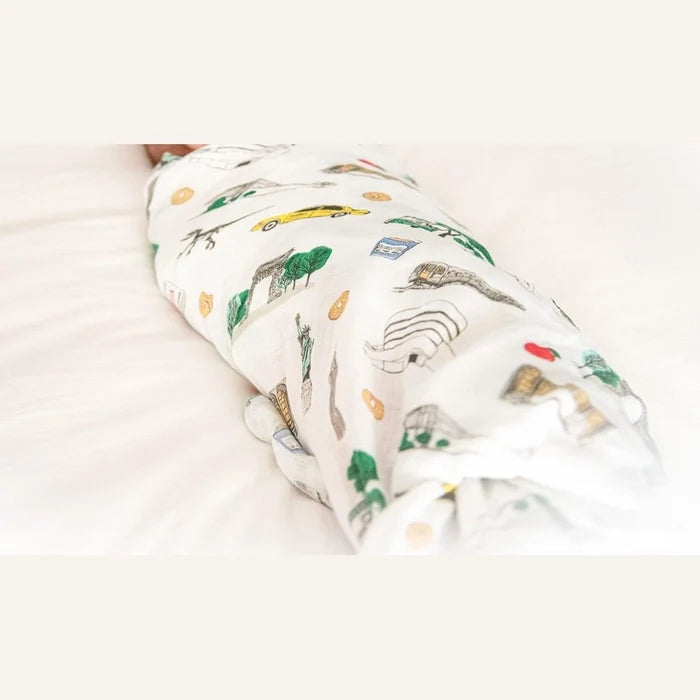 A baby swaddled in a white muslin blanket with a colorful NYC-themed print, lying on a white bed.