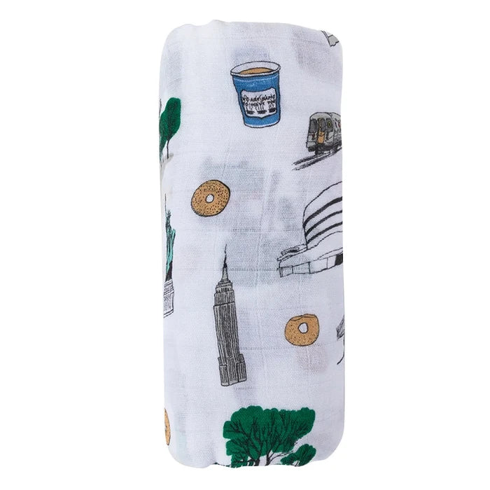 A rolled-up white muslin swaddle blanket featuring a colorful printed pattern of New York City landmarks, including the Empire State Building, Statue of the Liberty, Central Park, a bagel, and a blue Greek coffee cup.