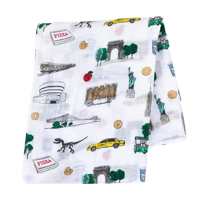 A white muslin baby blanket featuring a colorful NYC-themed print with illustrations of landmarks, taxis, a raptor, and pizza boxes.