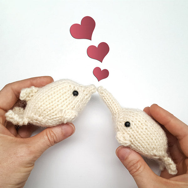 Two hands holding cream knitted dolphin and narwhal toys nose to nose with three pink hearts floating above them