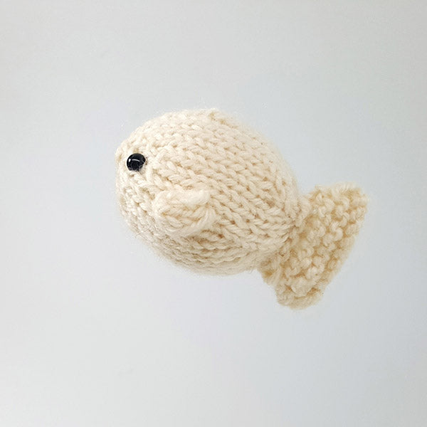 Close-up of cream knitted puffer fish toy with round body, small fins, and black bead eye on gray background