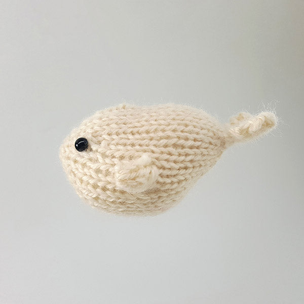 Close-up of cream knitted whale toy with elongated body, small curled tail, and black bead eye on gray background