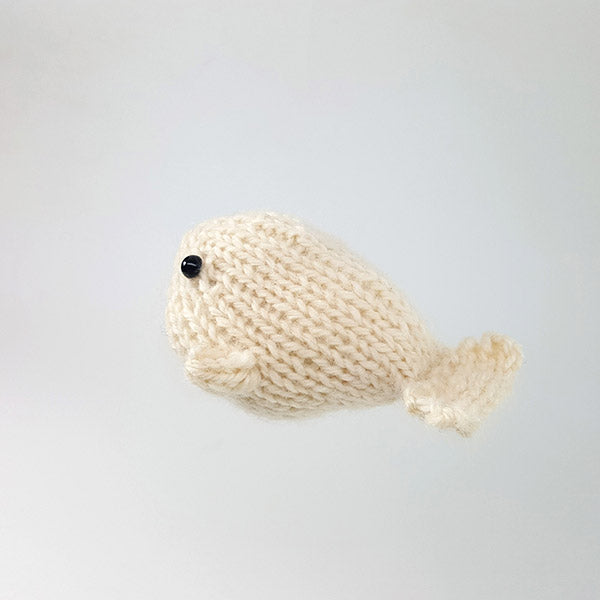 Profile view of cream knitted whale toy showing smooth stockinette body, small flipper, tail fin, and black bead eye on gray background