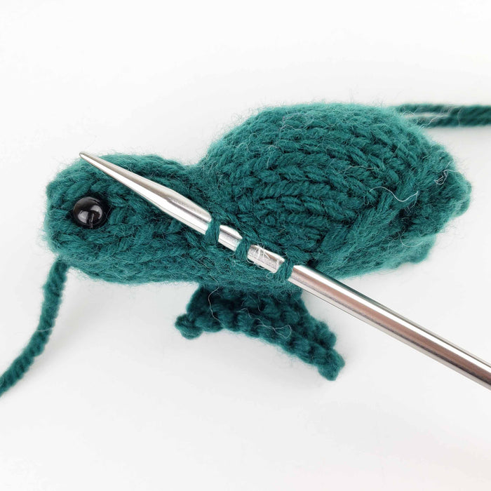 Close-up of teal knitted sea turtle in progress on double-pointed knitting needles with yarn tail visible, showing construction detail