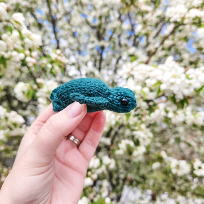 Hand holding a teal knitted sea turtle in profile against a background of white blossoming tree branches outdoors