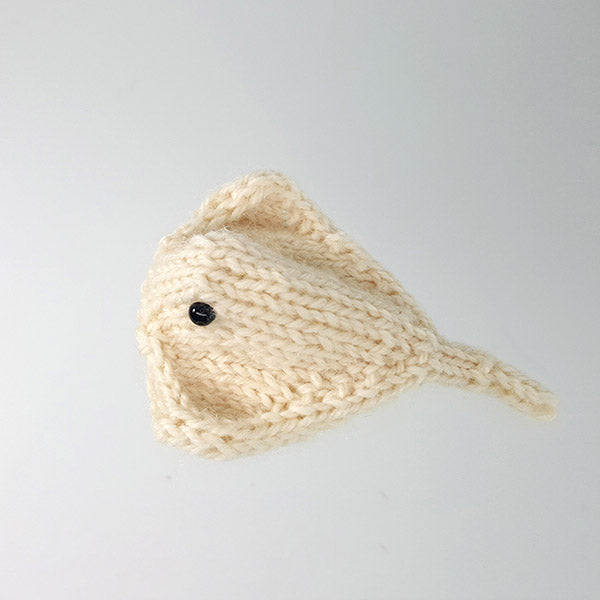 Cream knitted stingray toy with flat diamond-shaped body, long pointed tail, and black bead eye on gray background