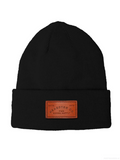 A black ribbed knit beanie hat with a folded cuff, featuring a brown rectangular leather patch on the front that reads 'HEADSTER CO. FUN GOODS SUPPLY'.