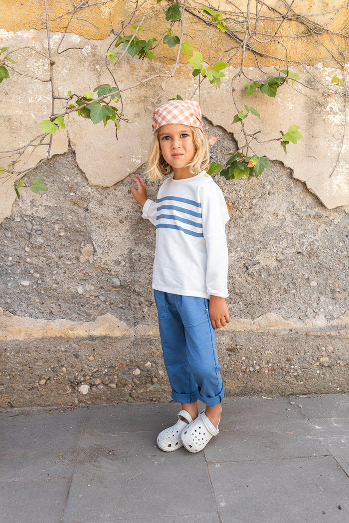 Child wearing a striped shirt and blue pants standing against a stone wall.