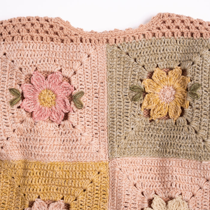 Close-up of a crocheted patchwork sweater with four pastel squares, each featuring a raised flower motif. The colors include blush pink, sage green, mustard yellow, and a warm beige/taupe.