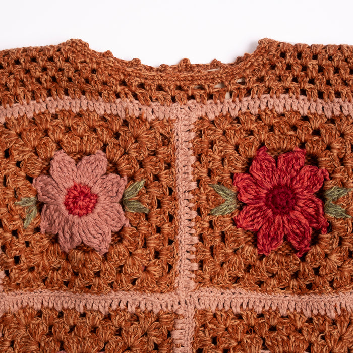 Close-up of a crocheted sweater with a warm rust-brown grid pattern and two embroidered flowers, one muted pink and one deep red, with small green leaf accents, set against a plain white background.