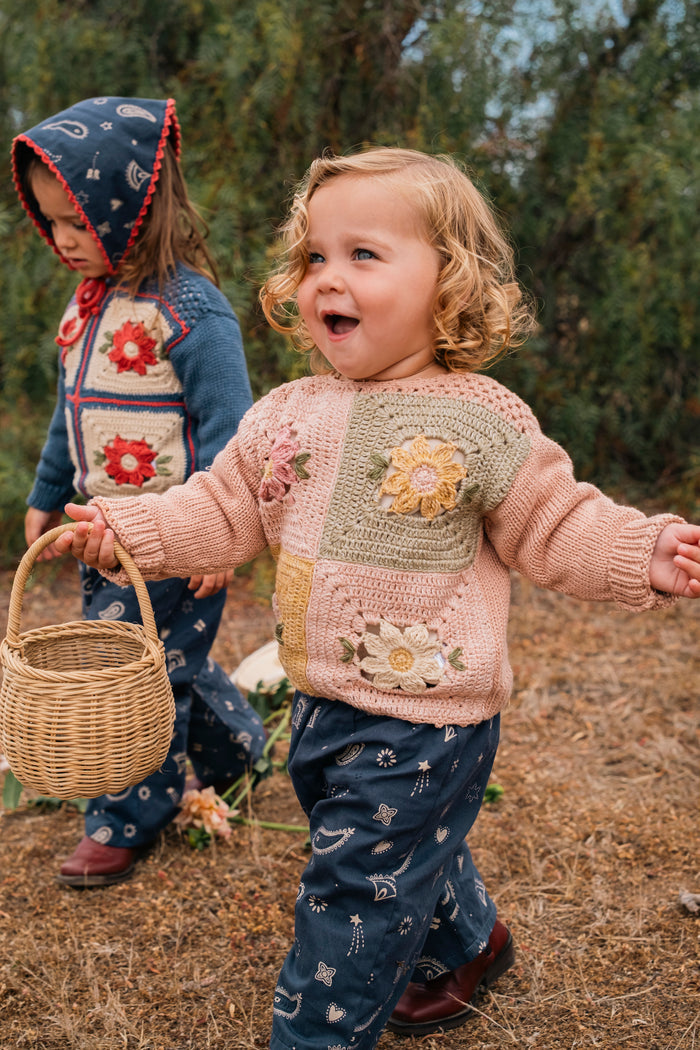 A toddler wearing a pink and beige crocheted patchwork sweater with floral details, blue patterned pants, and brown shoes walks outdoors in a field while holding a small woven basket. Another child is visible in the background wearing a blue hooded sweater.