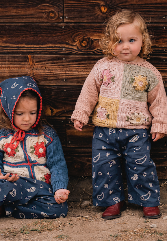 Two young children wearing handmade patchwork knit sweaters and patterned pants, posed in front of a rustic dark wood plank wall.
