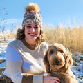 Woman wearing colorful crochet pawprint beanie with faux fur pom-pom, smiling outdoors with dog in snowy field