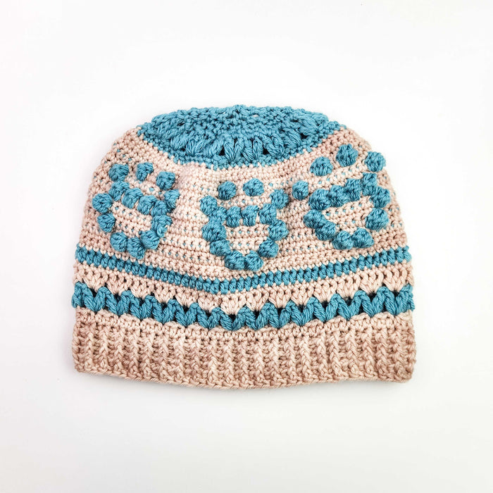 Blush and teal crochet beanie with raised pawprint bobble motifs laid flat on white background