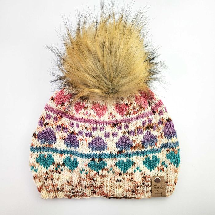 Colorwork knit beanie with pawprint motifs in pink, purple, and teal on cream with faux fur pom-pom