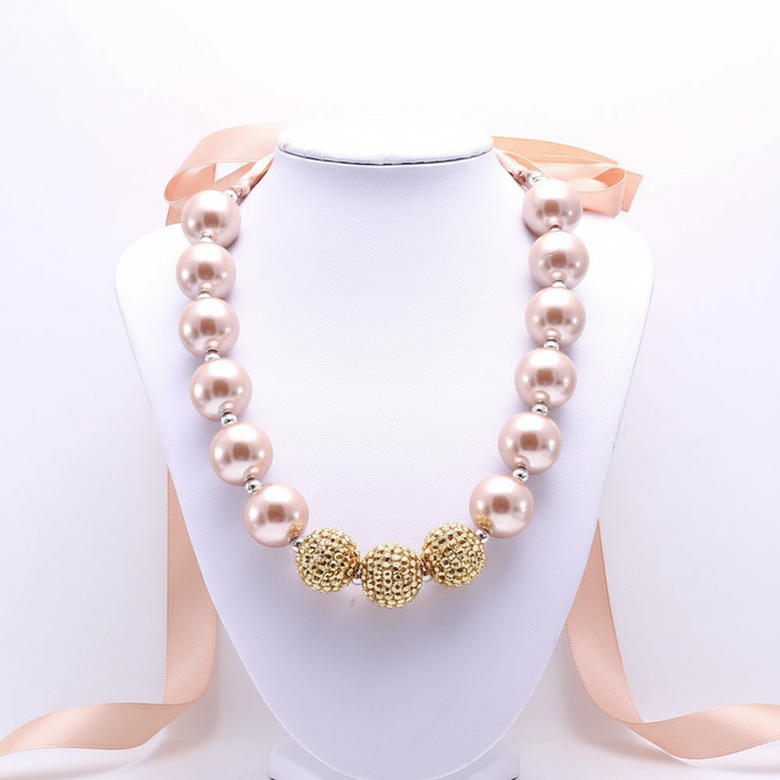A bold statement necklace featuring oversized blush-toned pearls and textured gold-finish focal beads, finished with a soft satin ribbon tie for a sophisticated, high-impact editorial look.