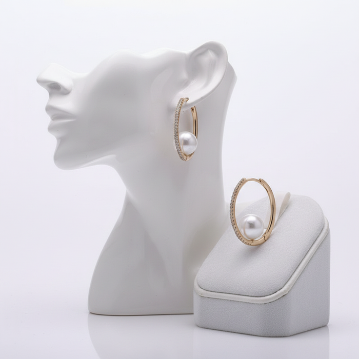 A pair of gold-toned oval hoop earrings featuring a shimmering pavé-set crystal front and a large, white "floating" pearl suspended within the bottom of each hoop. One earring is displayed on a white mannequin head, and the other is presented on a grey velvet jewelry stand, all against a clean white background.