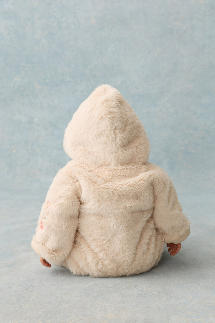back view of baby sitting on grey backdrop wearing cream faux fur romper with hood.