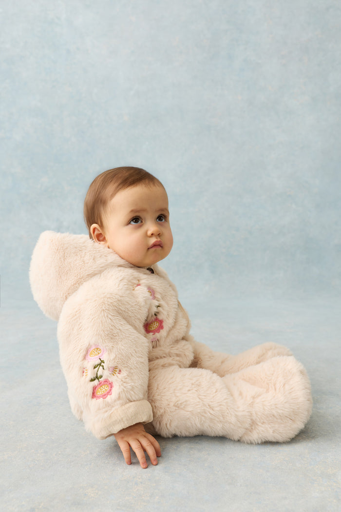 side view of baby sitting on grey backdrop wearing faux fur hooded romper with floral embroidery on sleeve and chest.