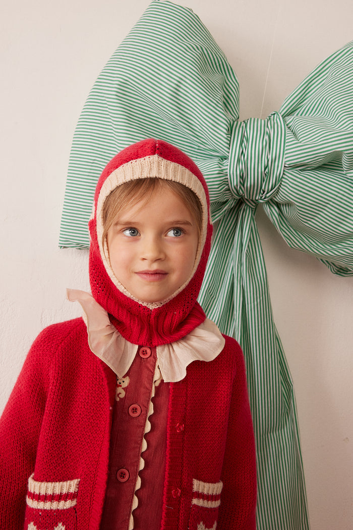 A young child wearing a festive red knit hooded cardigan with cream ruffle trim, posed in front of a large green and white striped bow on a light wall.