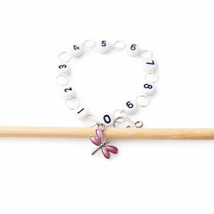 Pink dragonfly chain row counter bracelet with numbered white beads on a stretchy cord, draped over two wooden knitting needles
