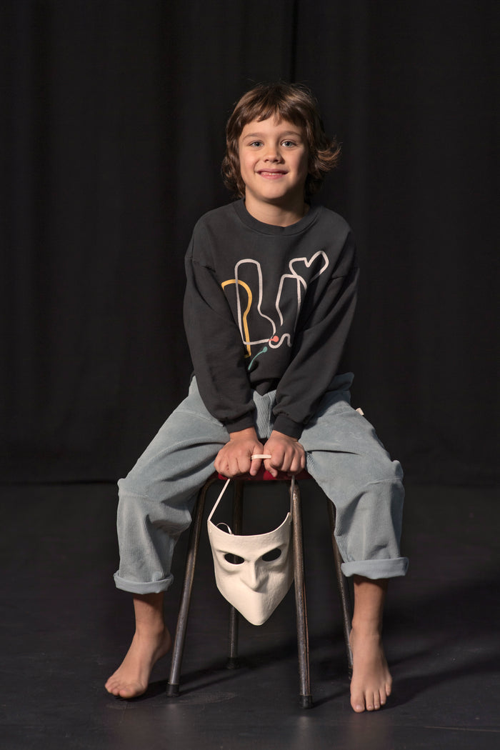 A young boy with short brown hair sits on a stool against a black curtain background. He is wearing a dark charcoal gray sweatshirt with a yellow and white graphic, light blue jeans, and is barefoot.