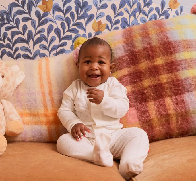 A smiling baby wearing a white long-sleeve footed romper sits on a soft tan couch in front of a large multicolor plaid cushion, with patterned wallpaper in the background.