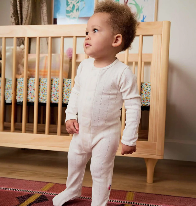 A baby wearing a cream ribbed long-sleeve footed sleeper stands in a nursery in front of a wooden crib.