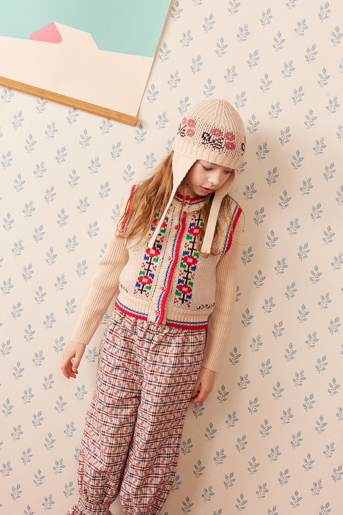 A young girl wearing a cream-colored knit hooded sweater vest with colorful floral embroidery, a matching cream knit beanie, and red-and-white plaid pants, standing against a patterned wallpaper background.