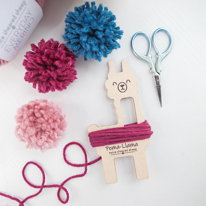 Poma-Llama tool with berry-colored yarn wrapped around its body surrounded by finished pom poms in pink, teal, and magenta, a skein of Everyday Merino yarn, and small scissors