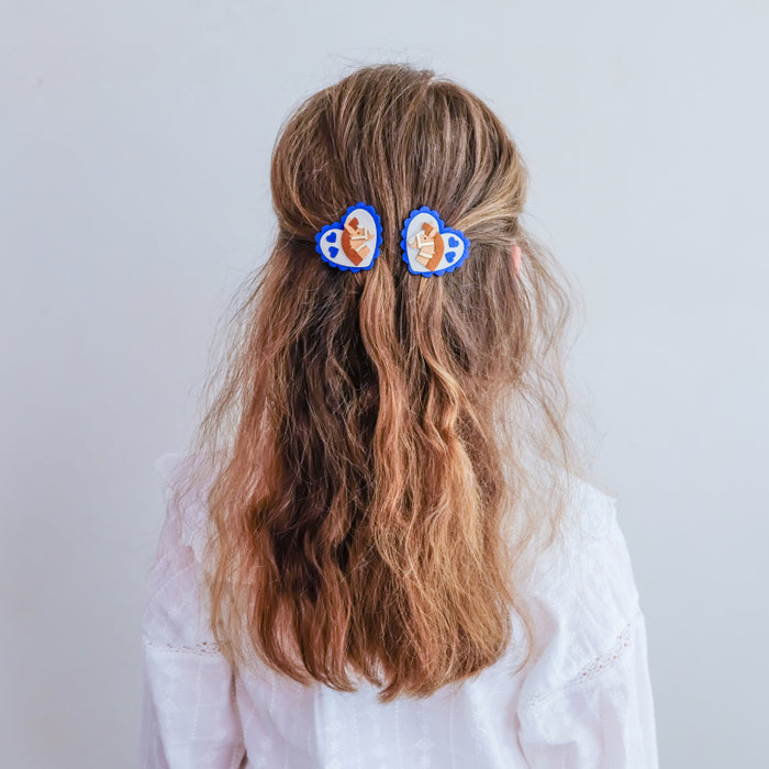 A person shown from the back with long wavy light brown hair, wearing two blue hair clips with cartoon faces, against a plain light gray background.